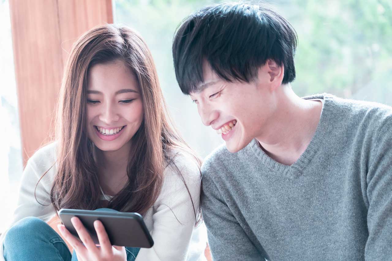 スマートフォンの画面に映るプロフィールムービーに夢中になっている笑顔の新郎新婦。