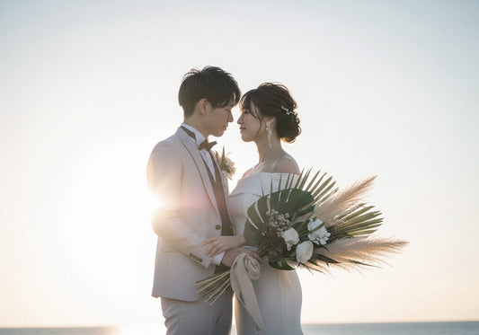 岩の多い海岸でロマンチックに抱き合う新郎新婦。花嫁はオフショルダーのドレスにブーケを持ち、新郎はベストと蝶ネクタイを締め、背後には夕日の黄金色の光が降り注いでいます。