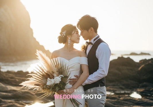 結婚式用の前撮りロケーション撮影をしている風景。岩の多い海岸でロマンチックに抱き合う新郎新婦。花嫁はオフショルダーのドレスにブーケを持ち、新郎はベストと蝶ネクタイを締め、背後には夕日の黄金色の光が降り注いでいます。
