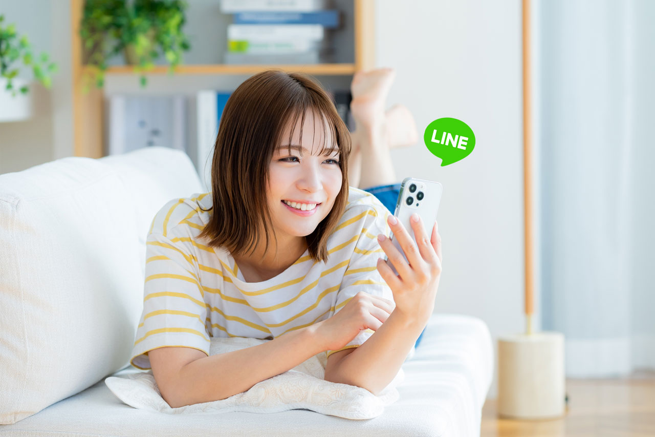 自宅でスマートフォンのLINEアプリを使い、プロフィールムービー作成用の写真を送信しながら笑顔を浮かべる若い女性。
